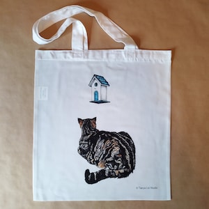 Puede incluir: Bolsa de tela blanca con un gato atigrado negro y marrón mirando una pequeña casita de pájaros blanca con un techo y una puerta azules. El texto "Tanya Lot Studio" está impreso en la esquina inferior derecha de la bolsa.