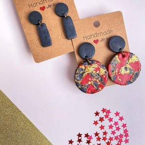 Könnte beinhalten: Handgefertigte Ohrringe auf braunen Karten. Ein Paar hat eine dunkelgraue Farbe mit einem kreisförmigen Oberteil und einem rechteckigen Boden. Das andere Paar hat ein rotes, schwarzes und goldenes kreisförmiges Design. Die Karten lesen "Handmade with love."