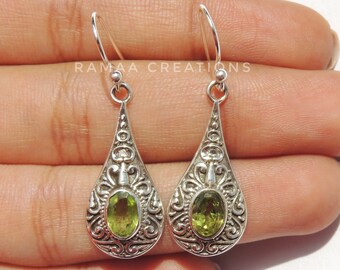 May include: A pair of silver dangle earrings with a green gemstone set in an ornate design. The earrings have a teardrop shape and are accented with intricate filigree details.