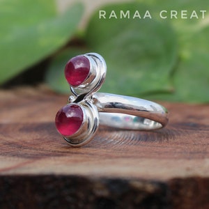 Pu&ograve; includere: Un anello d'argento con due pietre preziose rosa incastonate in un design a spirale. L'anello &egrave; su una superficie di legno.