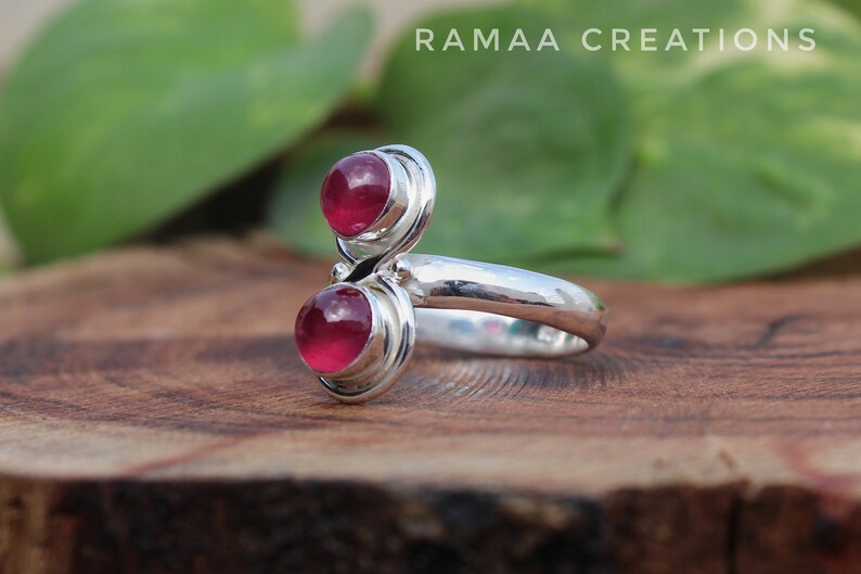 Pu&ograve; includere: Un anello d'argento con due pietre preziose rosa incastonate in un design a spirale. L'anello &egrave; su una superficie di legno.