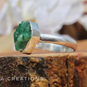 May include: A silver ring with a large, rough, green emerald gemstone. The ring is set on a wooden surface.