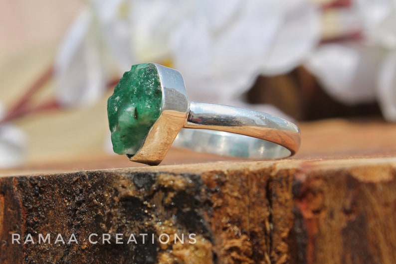 May include: A silver ring with a large, rough, green emerald gemstone. The ring is set on a wooden surface.