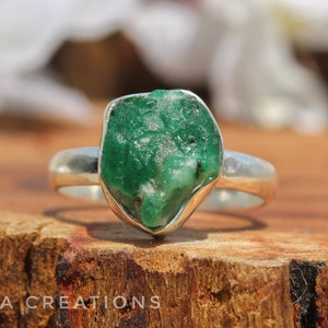 May include: A silver ring with a large, raw emerald gemstone. The emerald is a deep green color and has a rough, natural texture.
