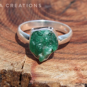 May include: A silver ring with a large, green, rough-cut emerald gemstone. The ring is set on a wooden surface.