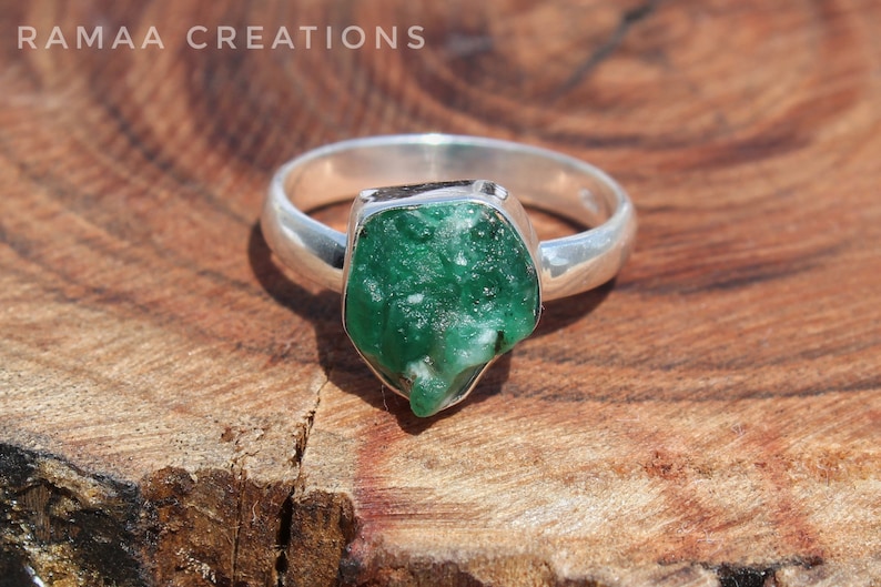 May include: A silver ring with a large, green, rough-cut emerald gemstone. The ring is set on a wooden surface.