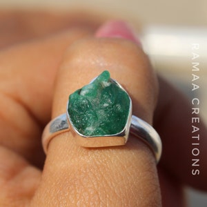 May include: A silver ring with a large, green, raw emerald gemstone set in the center. The ring is on a finger.