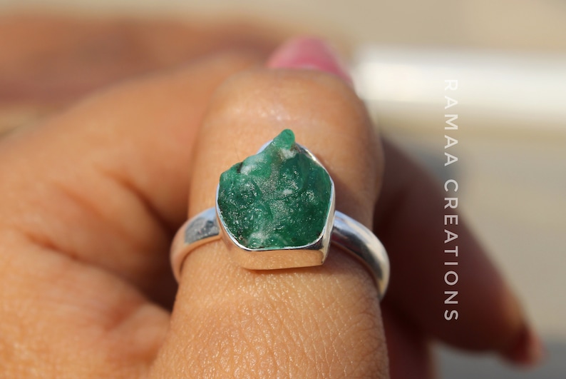 May include: A silver ring with a large, green, raw emerald gemstone set in the center. The ring is on a finger.