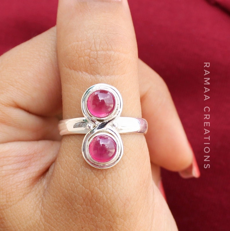 Pu&ograve; includere: Un anello d'argento con due pietre preziose rosa incastonate in un design a doppio infinito. L'anello &egrave; su un dito.