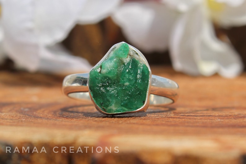 May include: A silver ring with a large, rough, green emerald gemstone. The ring is set on a wooden surface.