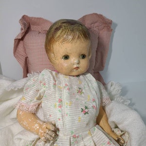 May include: A vintage porcelain doll with painted features, wearing a white dress with pink and yellow floral print. The doll is sitting in a white blanket.