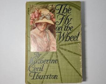 May include: A green book with the title "The Fly on the Wheel" by Katherine Cecil Thurston. The cover features a woman in a straw hat and a floral dress.