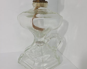 May include: A clear glass oil lamp with a brass burner and a handle. The lamp has a hexagonal base and a round chimney.
