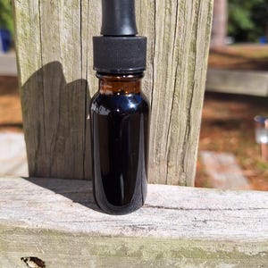 May include: A small, brown glass bottle with a black dropper top. The bottle is filled with a dark liquid. The bottle is sitting on a weathered wooden surface, with a wooden fence in the background.