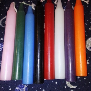 May include: Eight taper candles in various colors, including pink, green, blue, black, red, white, purple, and orange. The candles are arranged in a row on a dark blue background with a white star pattern.