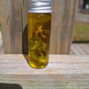 Puede incluir: Una pequeña botella de vidrio con tapa plateada llena de un líquido amarillo y materia vegetal verde y amarilla seca.