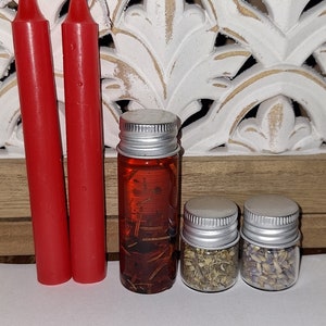 May include: Two red candles, a small bottle of brown liquid with herbs, and two small jars of dried herbs. The jars are labeled with white text.
