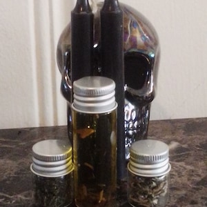 May include: Three small glass bottles with silver lids, two filled with dried herbs and one with a clear liquid, are arranged in front of two black candles and a black and iridescent skull.