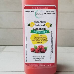 May include: A 473ml bottle of pink raspberry sea moss infused lemonade. The label reads "Sea Moss Infused Lemonade Raspberry" and includes the recommended daily serving of sea moss in every bottle. The bottle is on a white wooden surface.