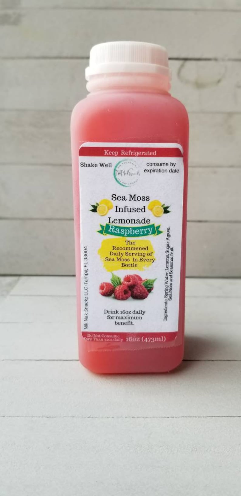 May include: A 473ml bottle of pink raspberry sea moss infused lemonade. The label reads "Sea Moss Infused Lemonade Raspberry" and includes the recommended daily serving of sea moss in every bottle. The bottle is on a white wooden surface.