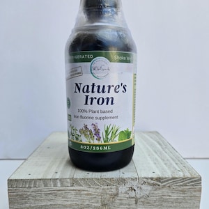 May include: A dark liquid bottle of Nature's Iron, a plant-based iron fluorine supplement, sits on a weathered wooden block. The label reads "Nature's Iron" and "8 oz/236 mL". The bottle is sealed with a clear plastic wrap.
