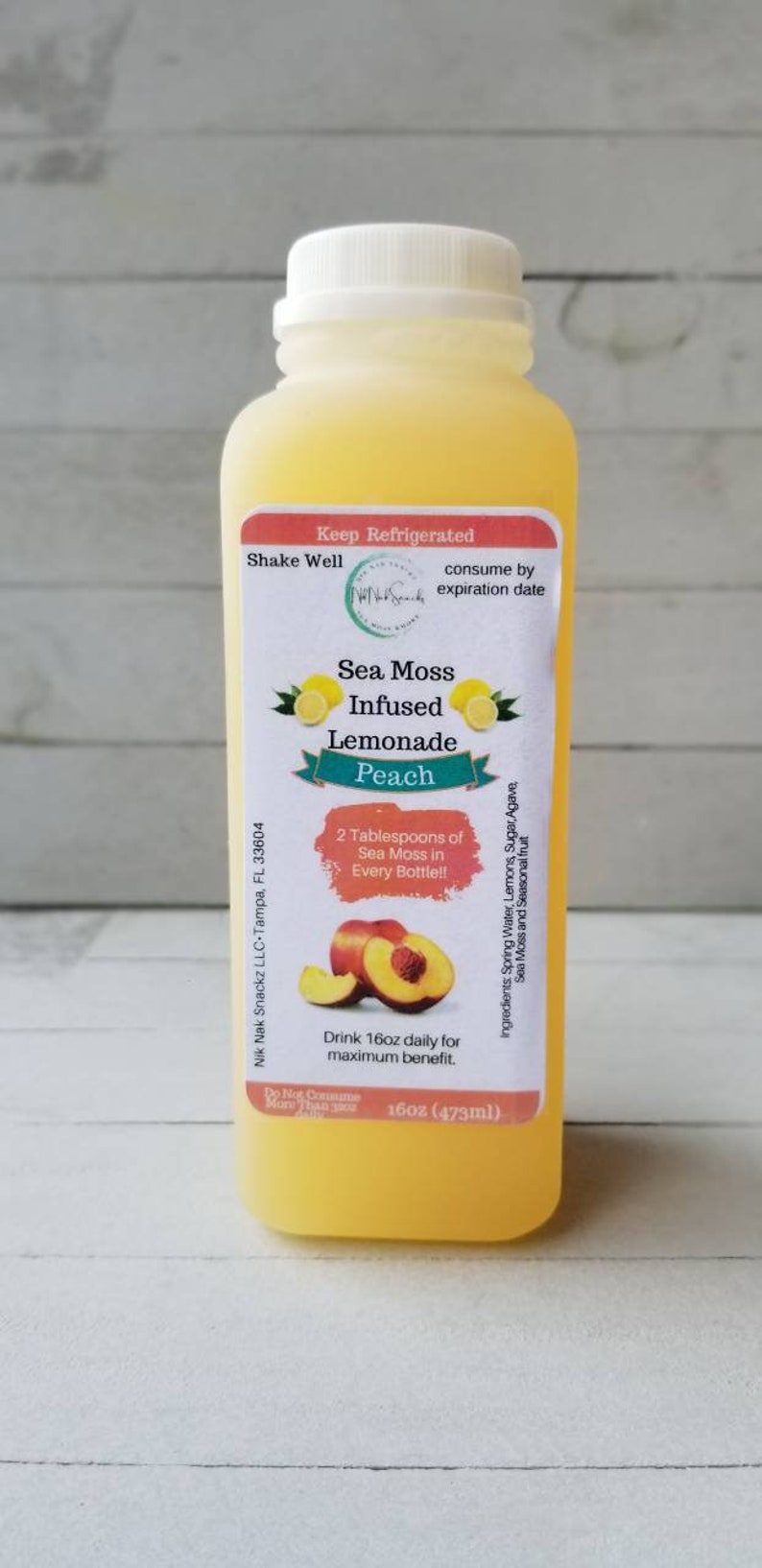 May include: A 473ml bottle of peach-flavoured sea moss infused lemonade. The label is white with a red border and features a peach slice and two lemons. The text on the label reads "Sea Moss Infused Lemonade Peach" and "2 Tablespoons of Sea Moss in Every Bottle!"