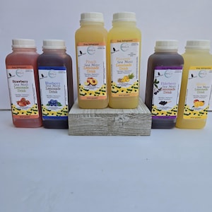 May include: Six glass bottles of sea moss lemonade drink in various flavours: strawberry, blueberry, peach, pineapple, elderberry, and mango. Each bottle has a label with the flavour and the words "Sea Moss Lemonade Drink".