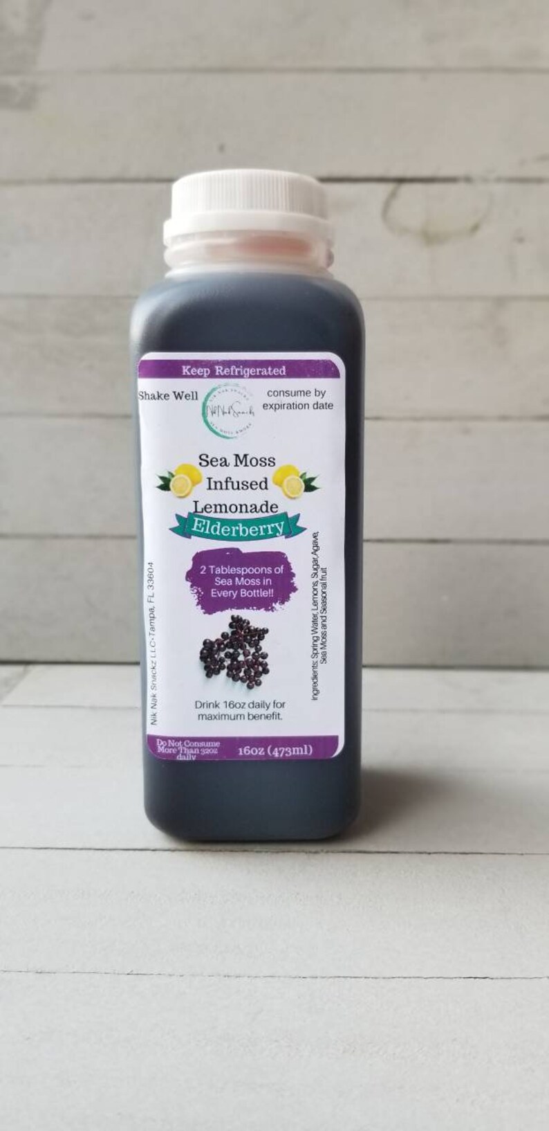 May include: A 473ml bottle of Sea Moss Infused Lemonade with Elderberry. The bottle is dark brown and has a white label with the product name, ingredients, and instructions. The label also features a picture of lemons and elderberries.