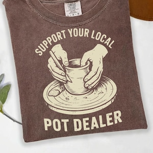 May include: Brown t-shirt with the text "SUPPORT YOUR LOCAL POT DEALER" in white. The graphic depicts hands shaping a pot on a pottery wheel. The shirt is a Comfort Colors brand.