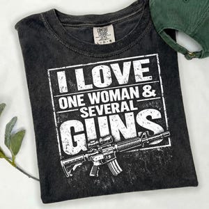 May include: Dark grey t-shirt featuring the text "I LOVE ONE WOMAN & SEVERAL GUNS" in a white rectangular design, with a rifle illustration. A green baseball cap is visible. The t-shirt is made of a soft material.