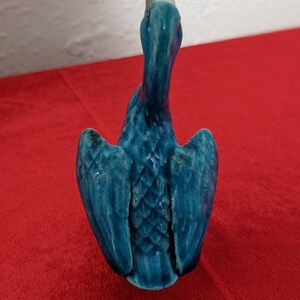 May include: A blue ceramic bird figurine with its wings spread out. The bird has a long neck and a small head. The figurine is sitting on a red surface.