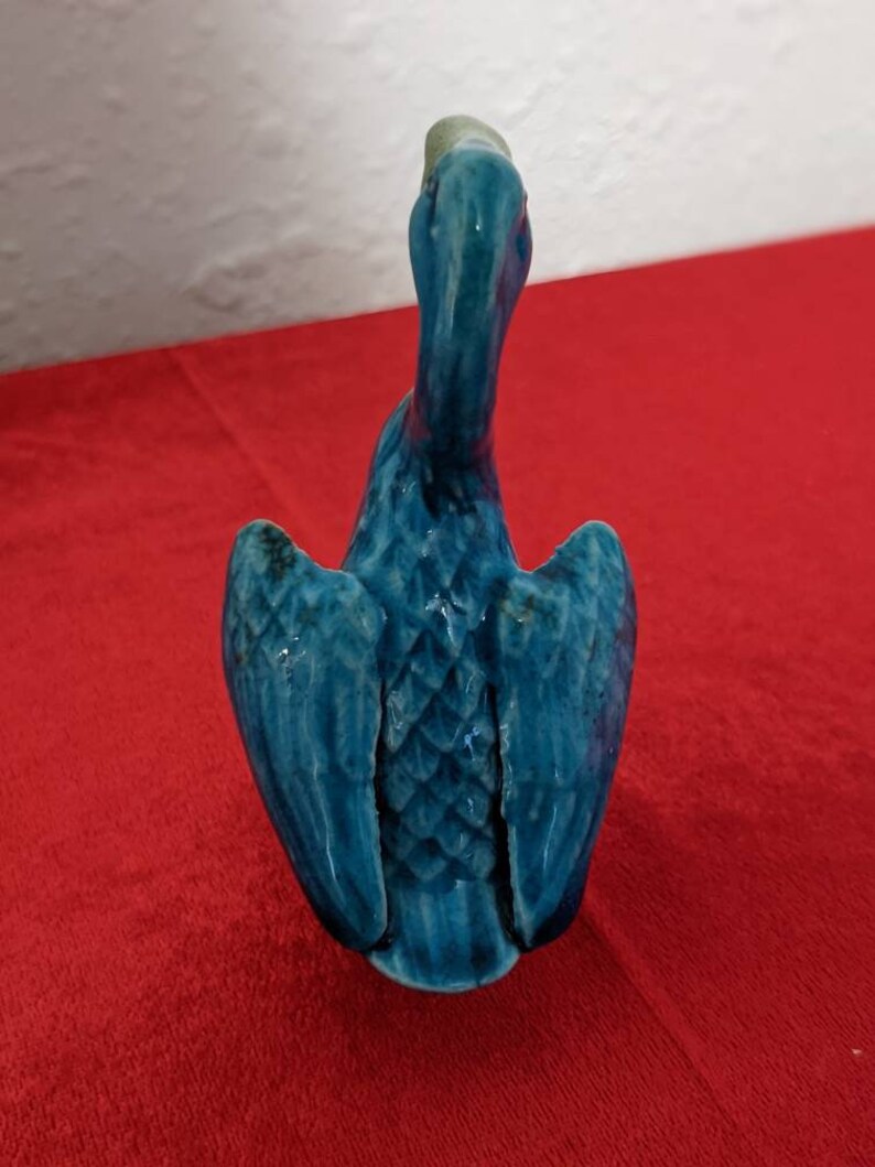 May include: A blue ceramic bird figurine with its wings spread out. The bird has a long neck and a small head. The figurine is sitting on a red surface.