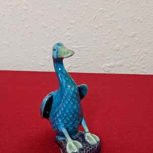 May include: A blue ceramic duck figurine with green accents. The duck is standing on a small, black base with a textured surface.
