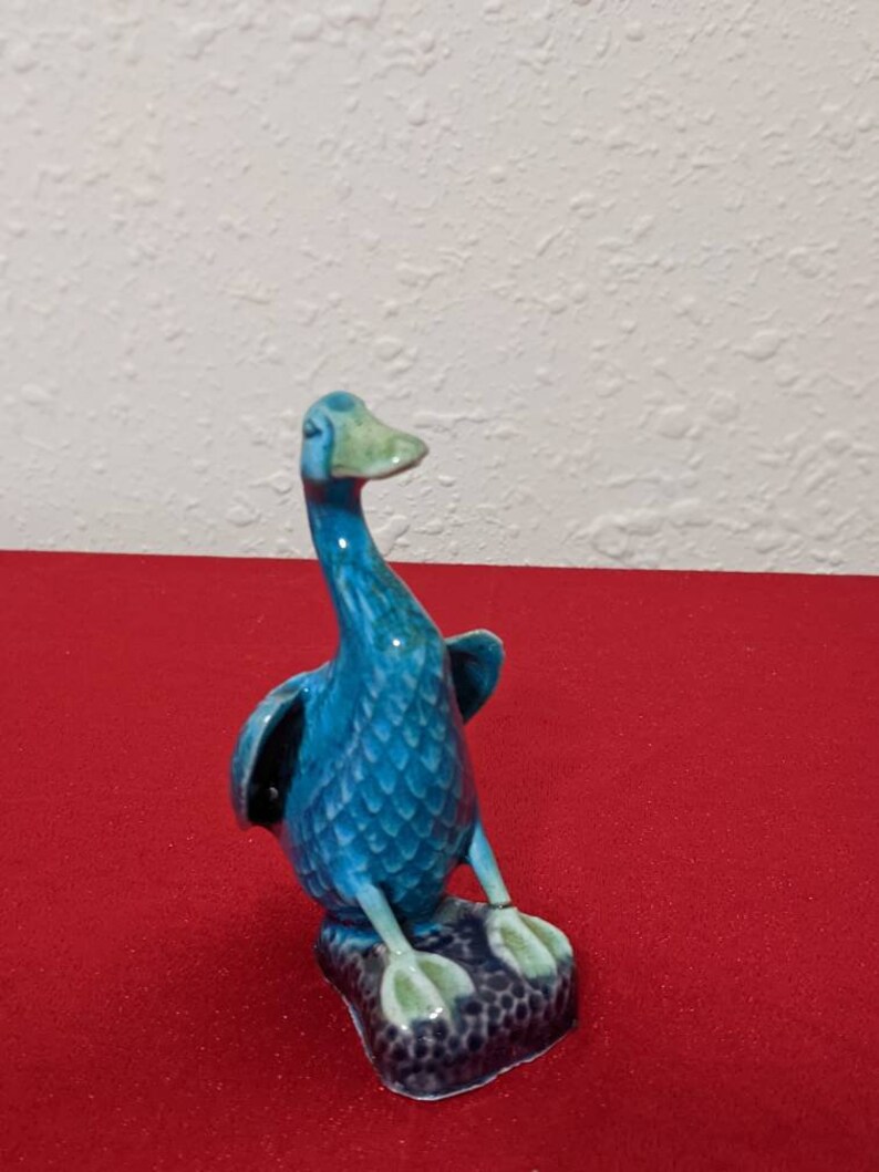 May include: A blue ceramic duck figurine with green accents. The duck is standing on a small, black base with a textured surface.
