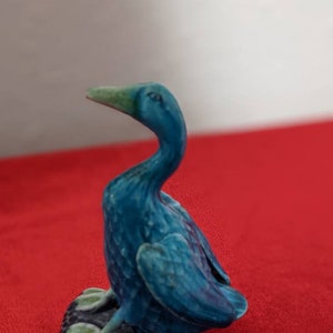 May include: A blue ceramic duck figurine with a green beak. The duck is sitting on a black base with green feet.