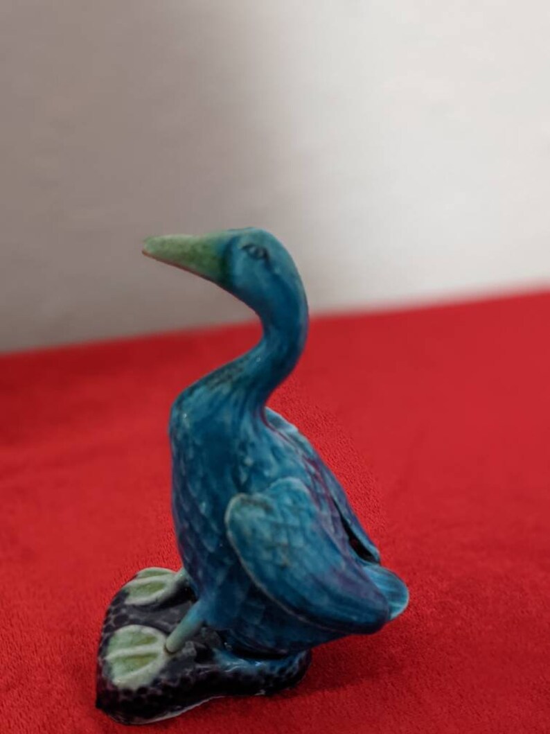 May include: A blue ceramic duck figurine with a green beak. The duck is sitting on a black base with green feet.