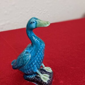 May include: A small blue ceramic duck figurine with a green beak and feet. The duck is sitting on a black base.