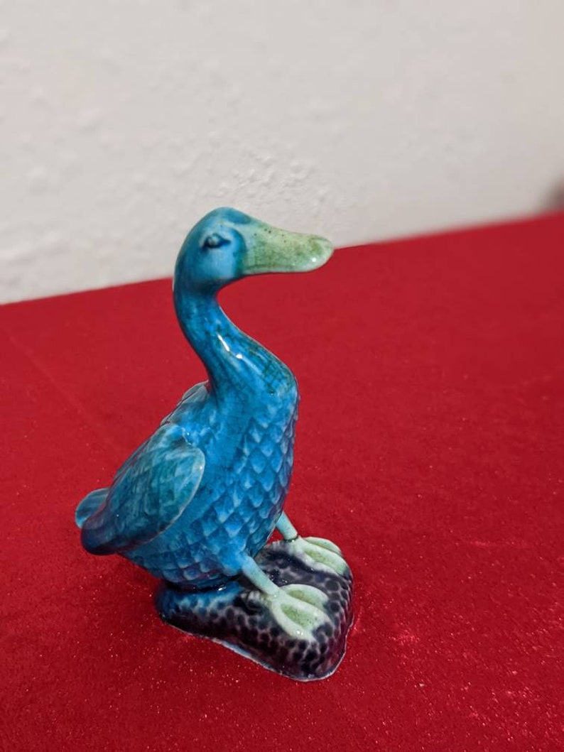 May include: A small blue ceramic duck figurine with a green beak and feet. The duck is sitting on a black base.