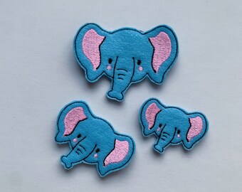 May include: Three blue elephant patches with pink inner ears and trunks are arranged on a white surface. The patches vary in size, with the largest at the top and two smaller ones below. Each elephant has embroidered details.