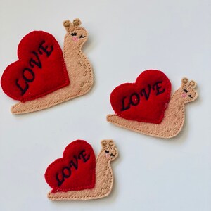 Puede incluir: Tres adornos de fieltro con forma de caracol y conchas rojas en forma de corazón. Cada corazón tiene la palabra "LOVE" bordada en negro. Los caracoles son de color beige con detalles cosidos, sobre un fondo blanco.