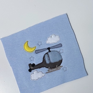 May include: A light blue fabric square featuring an embroidered design of a helicopter, clouds, stars, and a crescent moon. The helicopter is dark gray with a blue window. The moon is yellow, and the clouds are white.