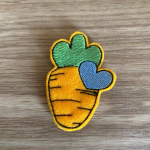 May include: A colorful embroidered patch featuring a carrot design with a green top and a blue heart. The carrot is orange with black details, and the heart is blue. The patch is likely for clothing or accessories.