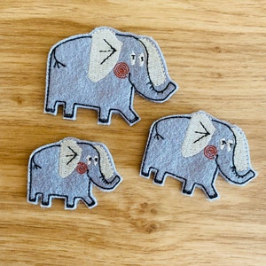 以下が含まれることがあります： クリーム色の耳と鼻を持つ、グレーのフェルト象のアップリケが3つ。頬には小さな赤い渦巻き模様があります。アップリケは黒で縁取られており、明るい茶色の木製の表面にあります。