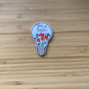 May include: White embroidered patch shaped like a light bulb with red and green flowers inside and the text "Be a light" on the top.
