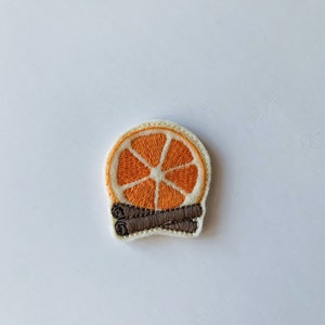 May include: Embroidered patch featuring a slice of orange with a white rind and a brown cinnamon stick. The patch has a cream-colored border and is set against a white background. The design is detailed and colorful.