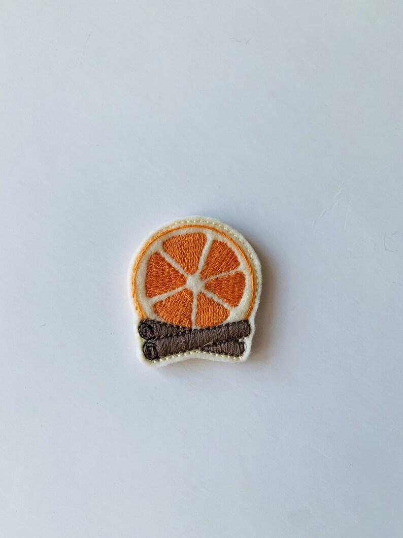 May include: Embroidered patch featuring a slice of orange with a white rind and a brown cinnamon stick. The patch has a cream-colored border and is set against a white background. The design is detailed and colorful.