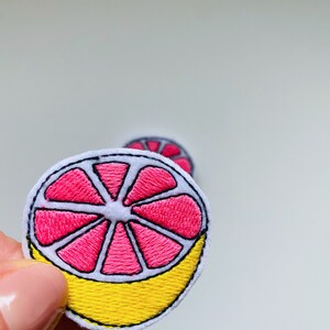 Op de afbeelding: Geborduurde patch met een roze grapefruit schijf met een witte schil en gele schil. De patch heeft een gedetailleerd gestikt ontwerp, met de nadruk op de segmenten van de vrucht. De patch is rond en heeft een witte rand.