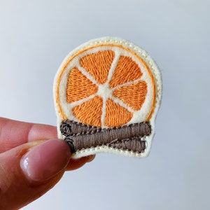 Puede incluir: Parche bordado con una rodaja de naranja y palitos de canela. La naranja es de color naranja brillante, con detalles blancos y crema. El parche está diseñado para ropa o accesorios.