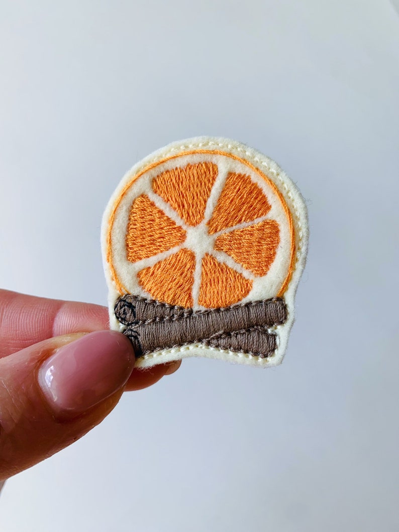 May include: Embroidered patch featuring a slice of orange with brown cinnamon sticks. The orange slice is bright orange with white and cream-colored details. The patch is likely for clothing or accessories.