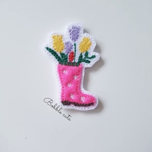May include: A pink rain boot with white polka dots filled with yellow and purple flowers. The boot is made of felt and has a white background.